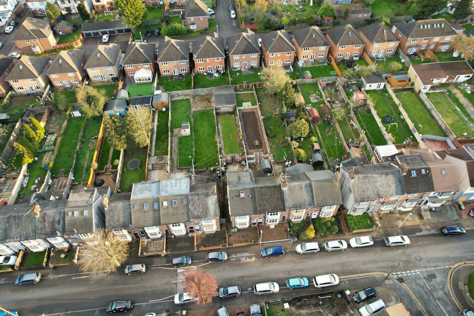 An aerial view of a suburban residential street showcasing a row of terraced houses with individual back gardens. The gardens vary in layout and maintenance, featuring lush green lawns, flower beds, p