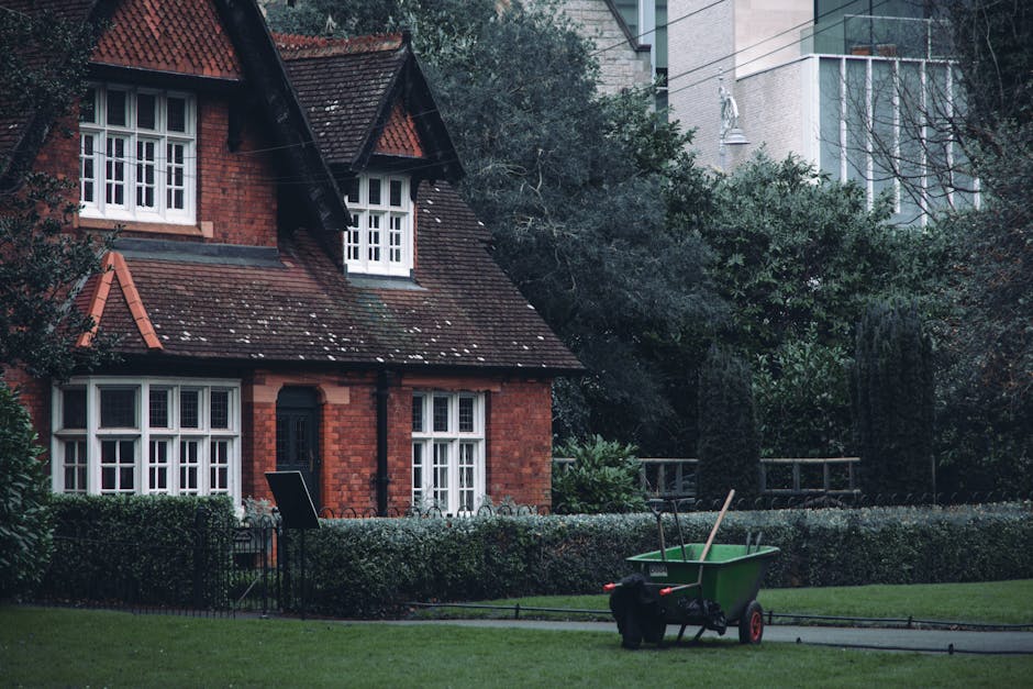 A well-maintained suburban garden in front of a traditional red brick house with white-framed windows and a sloped tiled roof. The garden features a neatly trimmed hedge bordering a lush, green lawn w