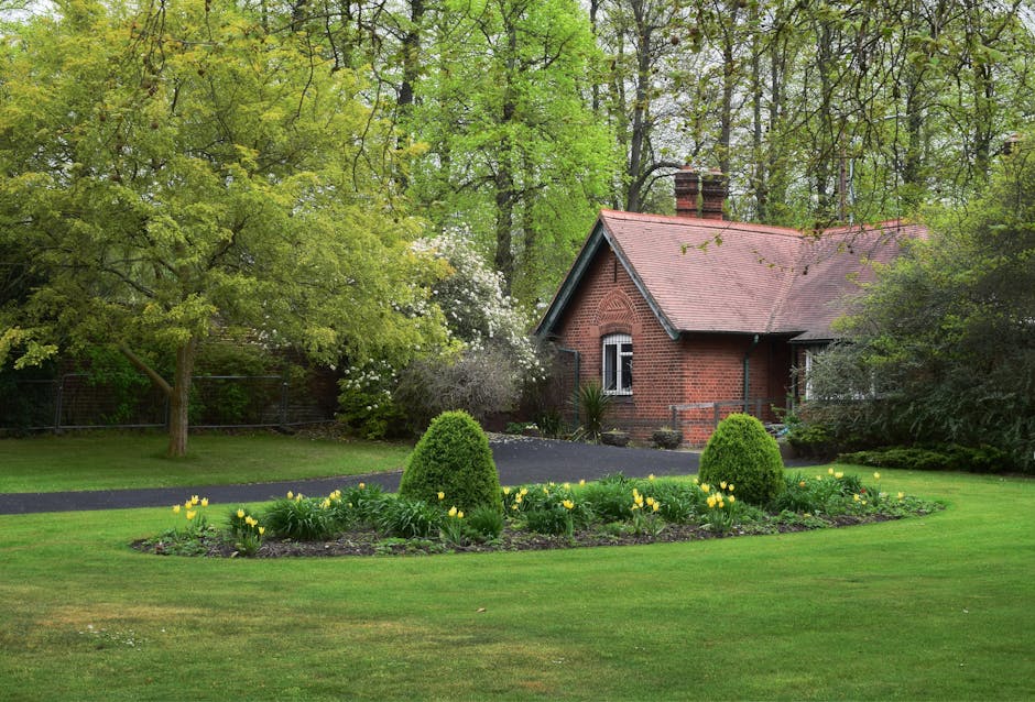 A well-maintained suburban garden in front of a traditional red brick house with white-framed windows and a sloped tiled roof. The garden features a neatly trimmed hedge bordering a lush, green lawn w